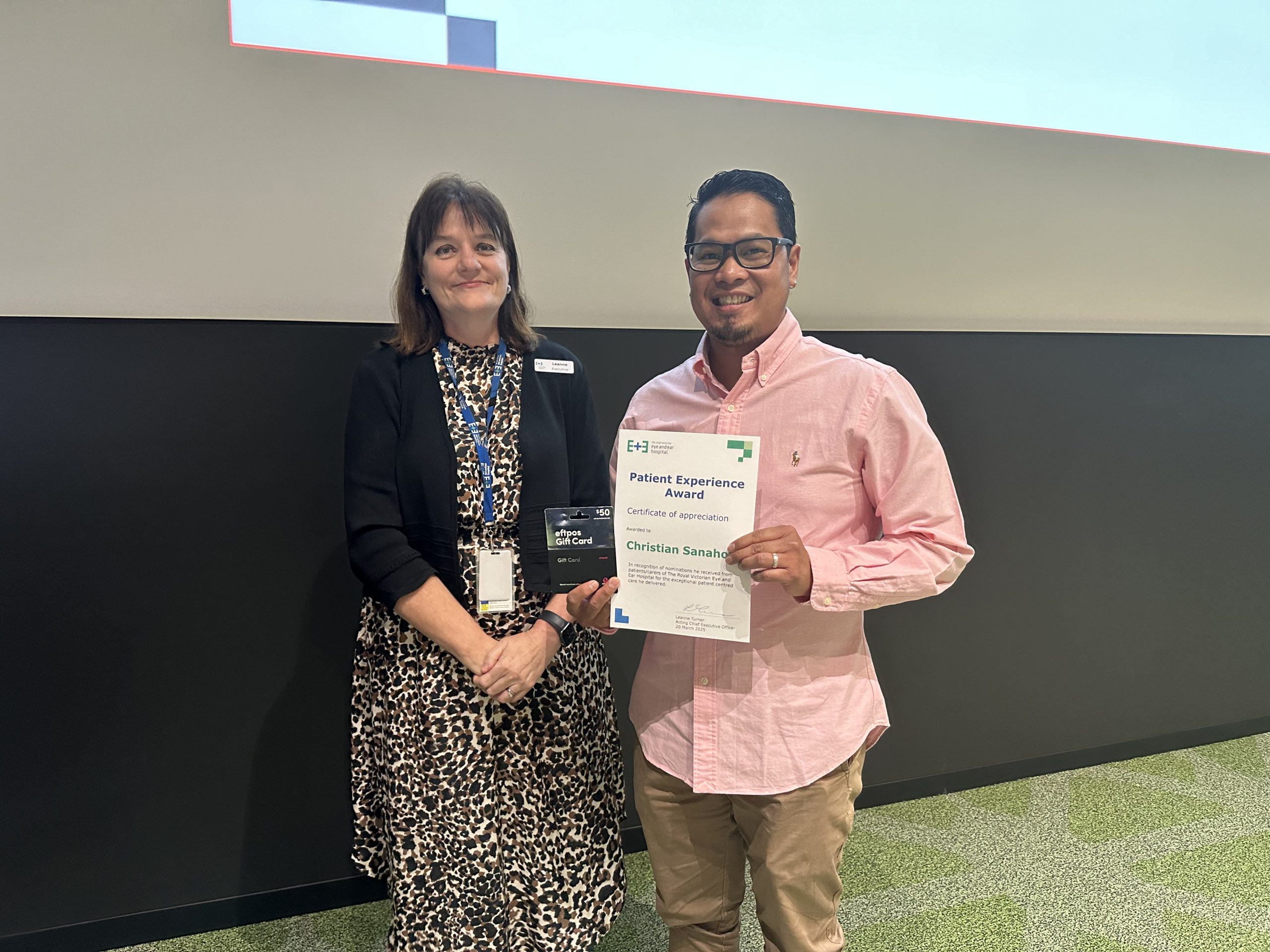 A man in a pink shirt, holding a certificate, standing next to a woman in a patterned dress and black blazer.