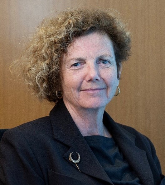A woman with light brown curly hair smiles at the camera against a brown paneled background wearing a black top and blazer