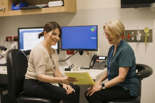 Two women sitting together in a consult room with a computer in the background. A woman with dark her is holding a yellow patient file towards a woman with short blonde hair. They are both smiling.