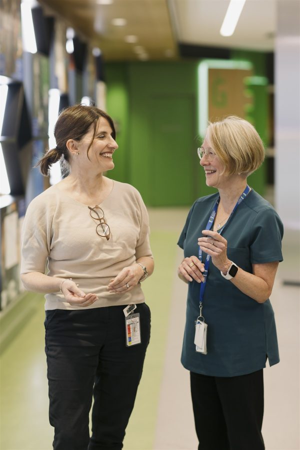 A woman with brown hair tied back in a ponytail wearing a cream top stands next to a woman with shorter blonde hair in teal medical scrubs against a green background.