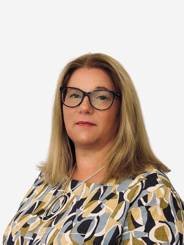 A blonde haired woman with black framed glasses smiles at the camera. She wears an olive, black, blue and white patterned blouse.