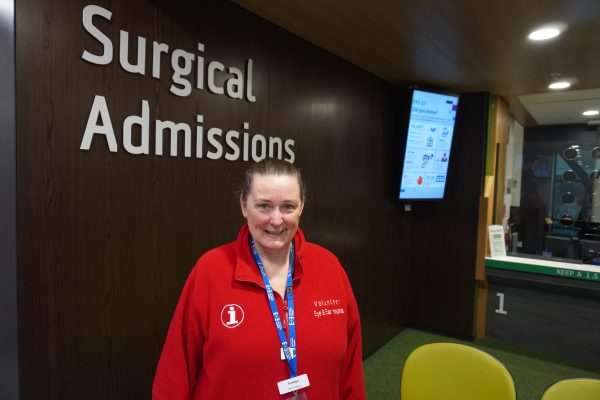 Teresa standing in front of the Surgical Admissions sign in the signature red volunteering jacket. She smiles at the camera.