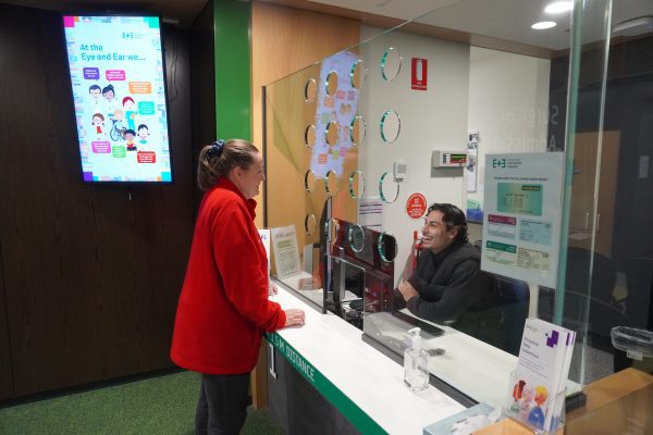 Teresa standing at the admissions desk chatting with one of the clerical staff who is behind a perspex screen. Both are smiling at each other. there is a Digital Display Screen in the background.