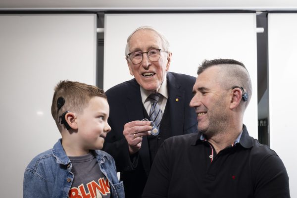 A young boy with a cochlear implant in a blue shirt, left, with Professor Clark holding a cochlear implant in a black suit, and a man in a black polo (right) with a cochlear implant