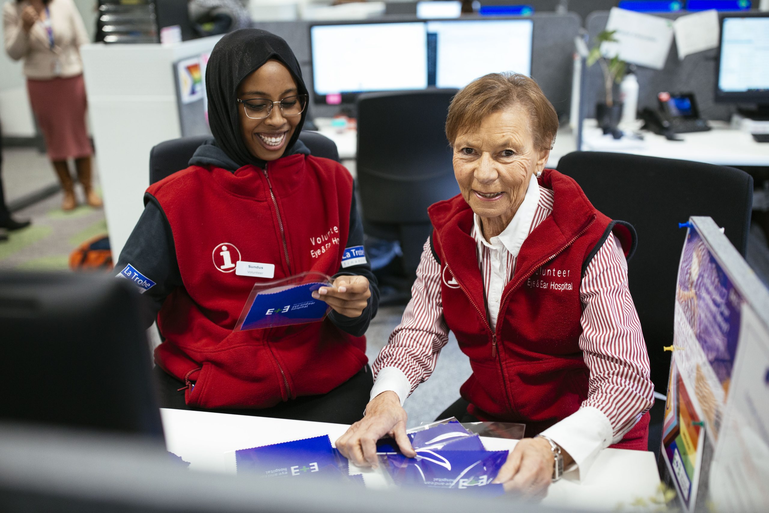Two volunteers wearing red vests sitting at a desk and smiling.