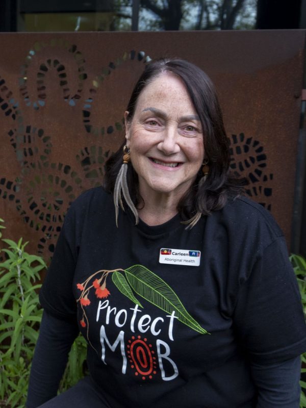 A woman with brown hair wearing emu feather earrings and a black t-shirt that says 'protect mob' smiles at the camera.