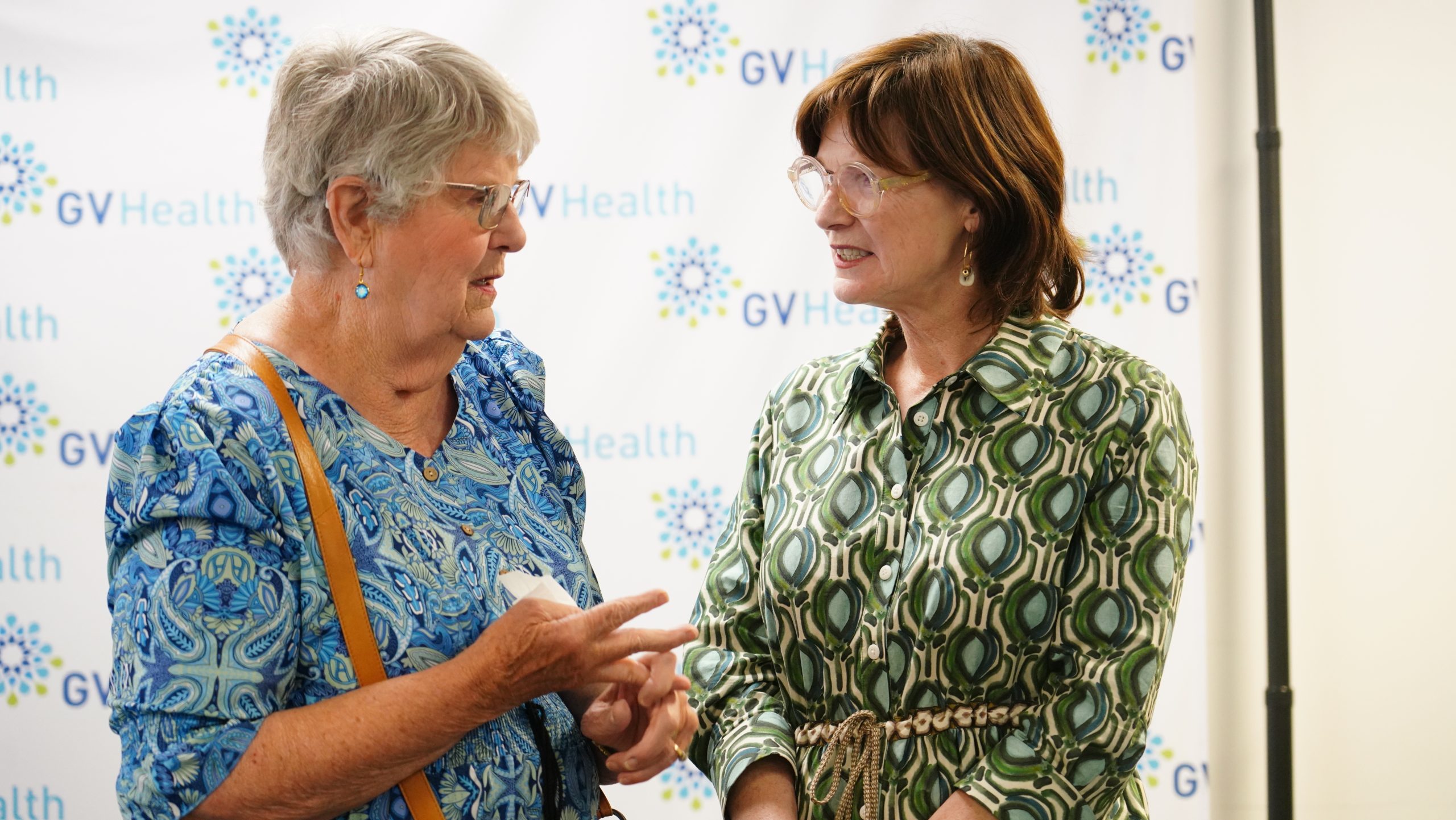 A woman in a green and white dress stands talking to an older woman in a blue and white dress.