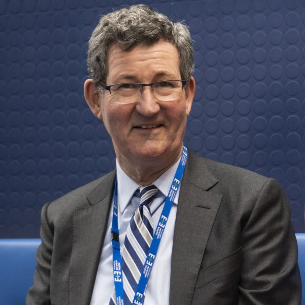 A photo of a man in a pale blue shirt and charcoal coloured suit jacket smiling at the camera. He wears glasses and an Eye and Ear lanyard around his neck.