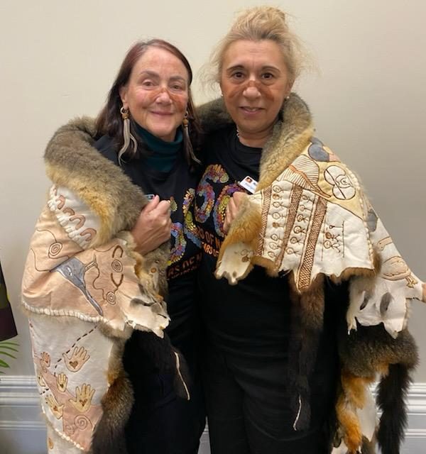 An image of two women draped in the Eye and Ear possum skin cloak, smiling at the camera with face paint across their noses.