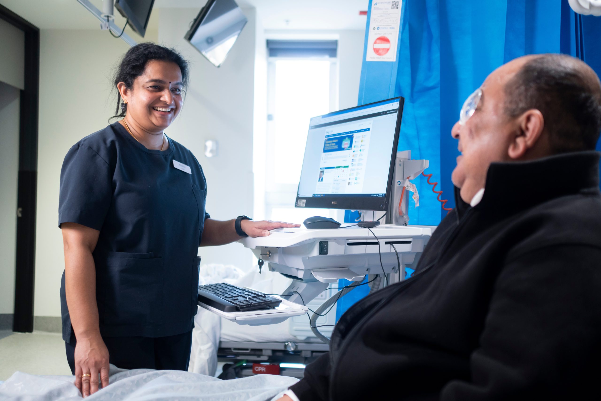 An inpatient nurse standing at a computer smiles down at a patient lying in a bed with an eye patch on.