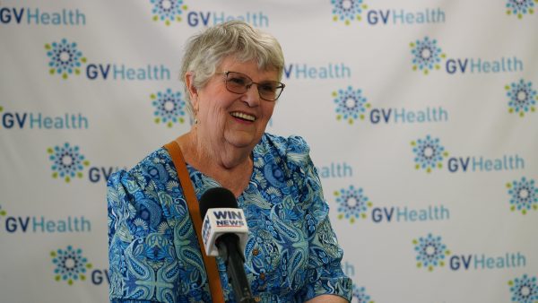 A smiling woman with glasses in a blue patterned dress speaking into a microphone.