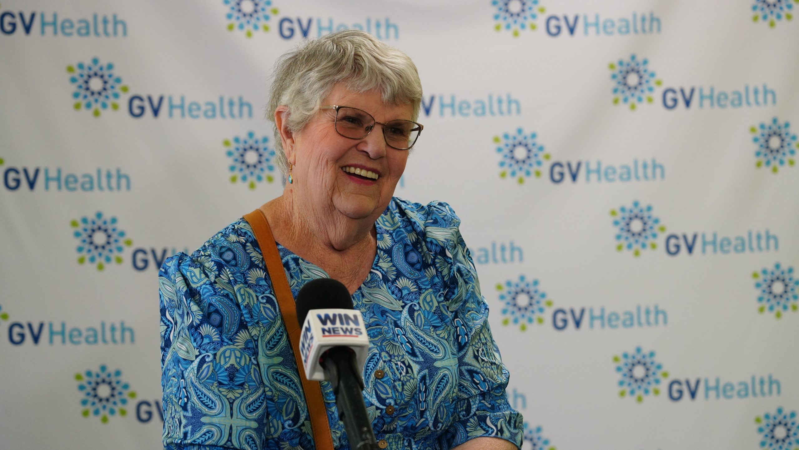 A smiling woman with glasses in a blue patterned dress speaking into a microphone.