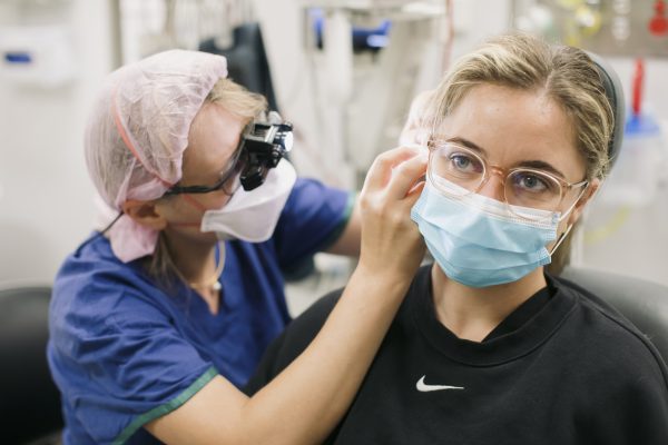 A doctor wearing blue scrubs and a mask looks inside a patients ear.