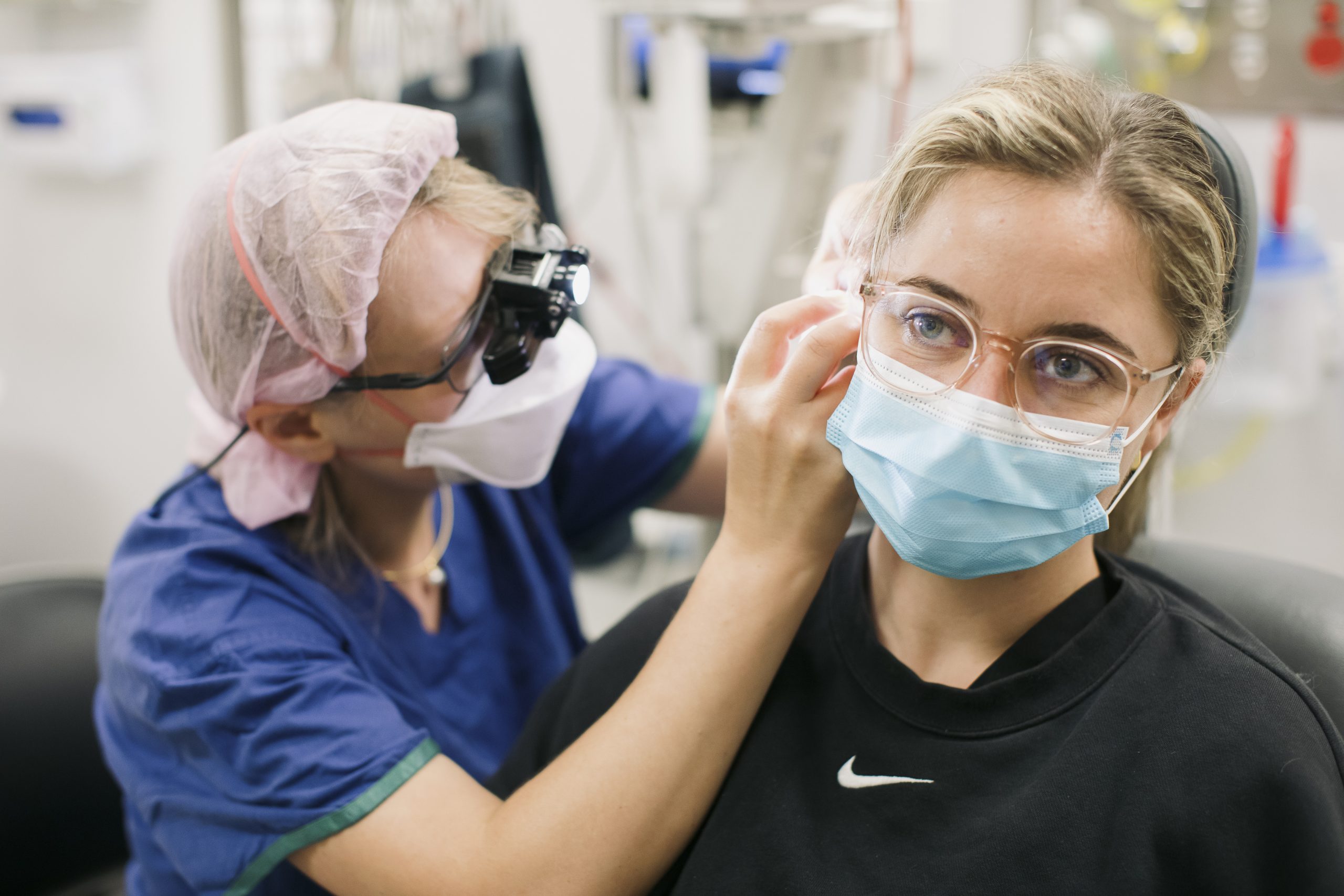 A doctor wearing blue scrubs and a mask looks inside a patients ear.