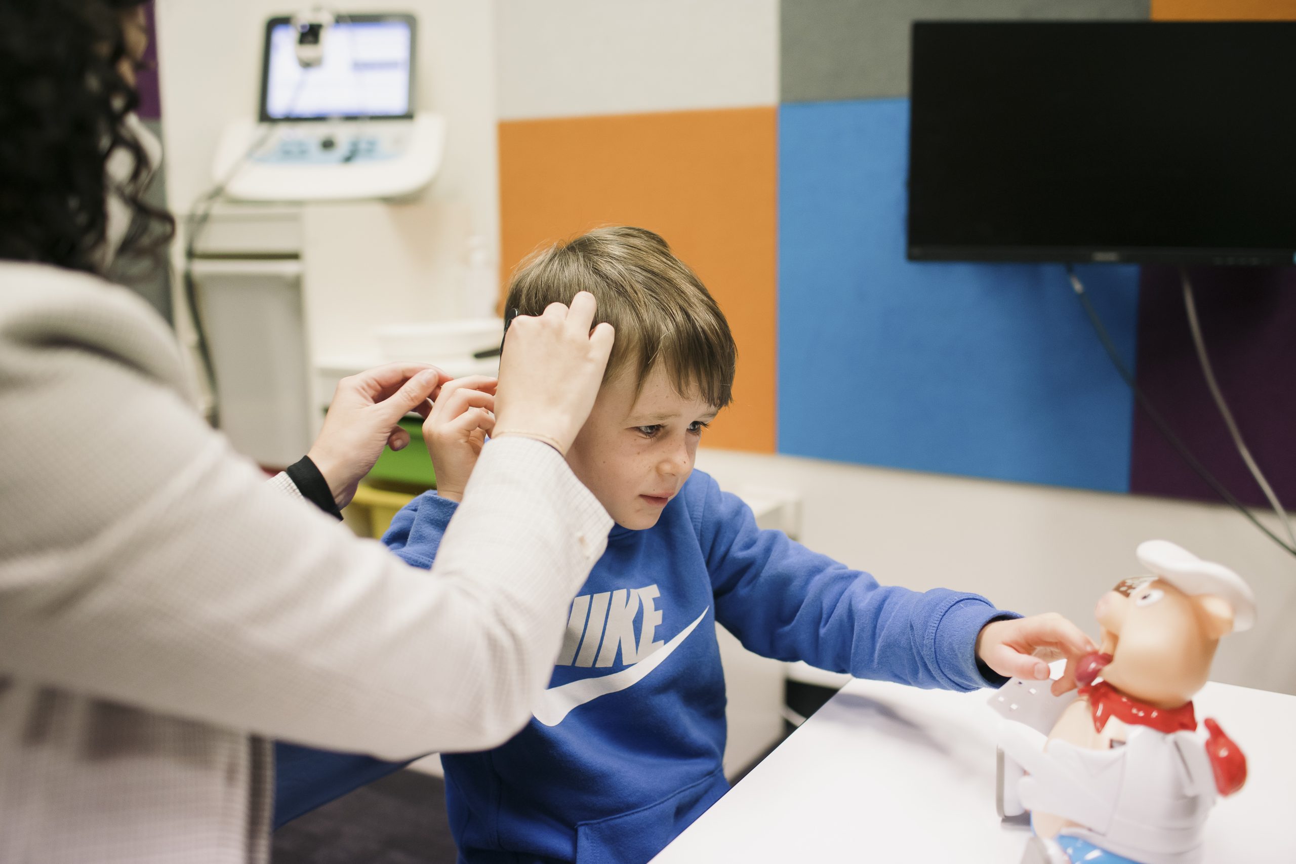 A photo of a young boy in a blue jumper having his cochlear implant adjusted by his speech pathologist.