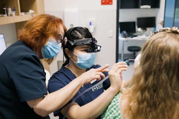 A nurse with red hair is instructing a younger nurse on how to use ear tools on a patient