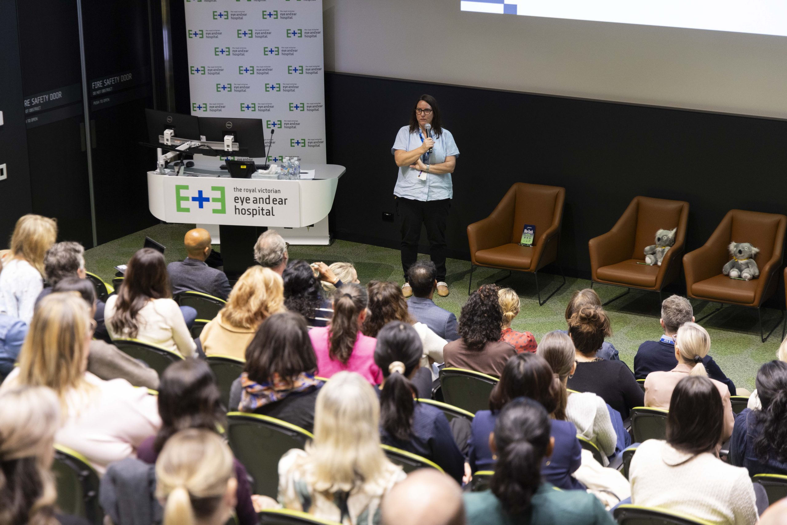 A woman in a light blue top holding a microphone and speaking to people in an auditorium.