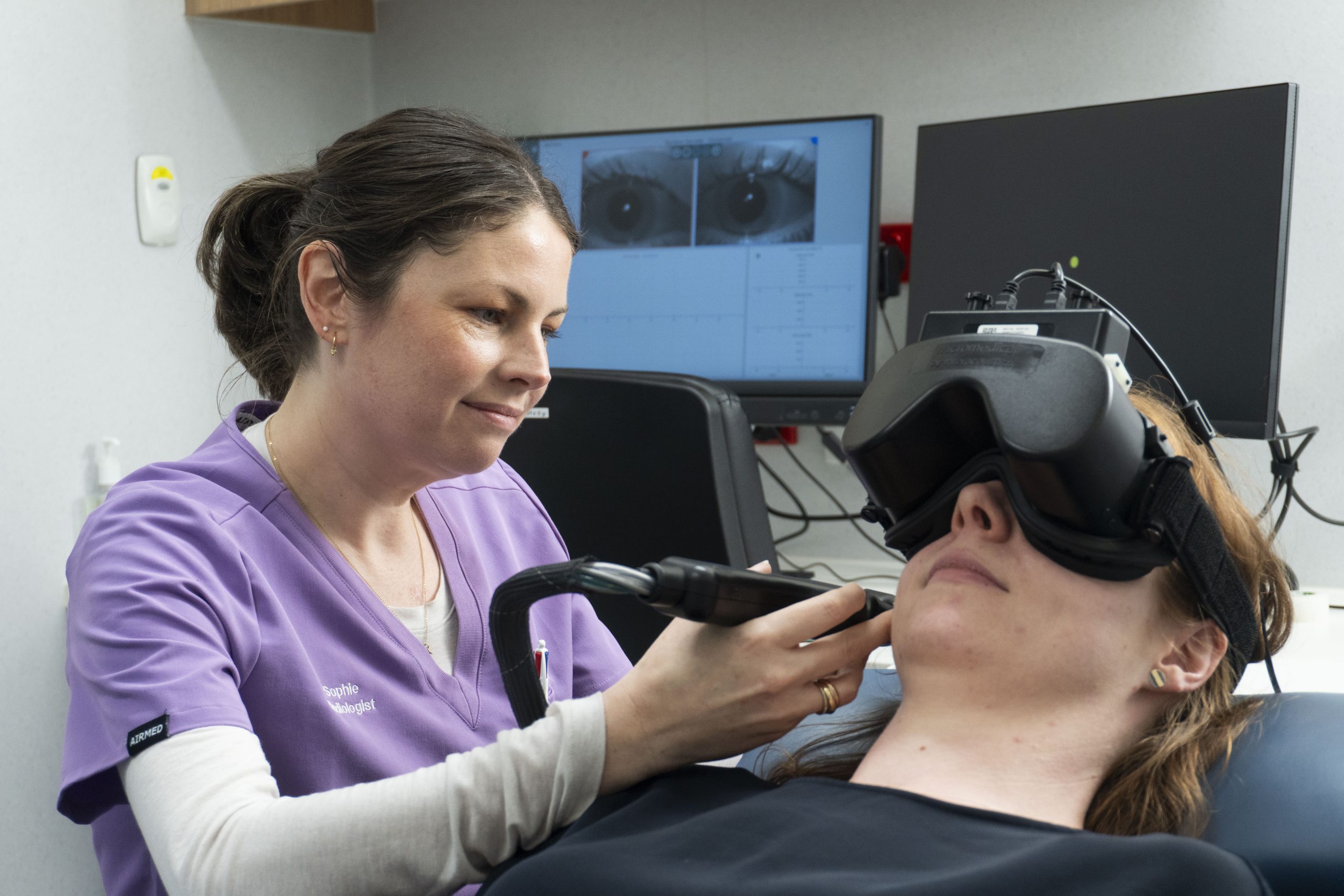 A woman lying on a chair wear large black goggles whilst a clinician in a purple scrub top holds an instrument.