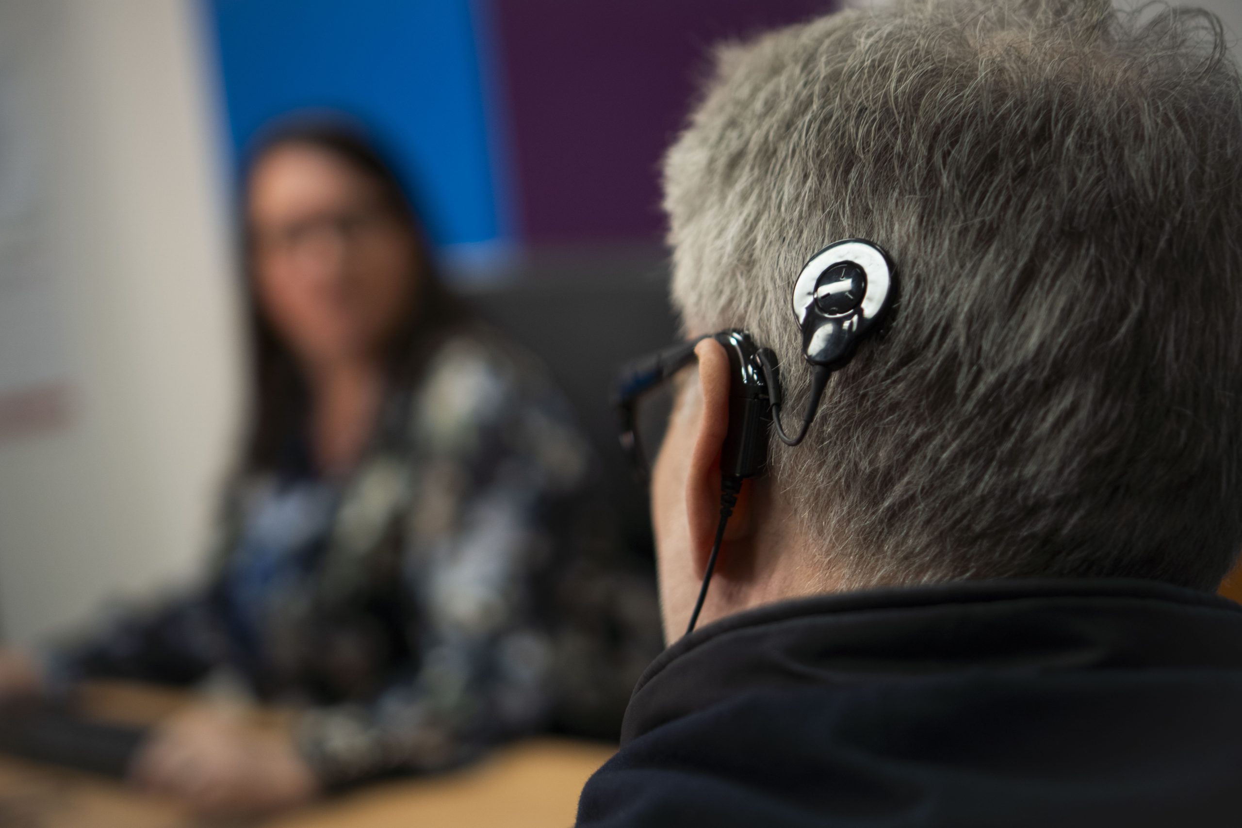 A close up of a cochlear implant on a head with a woman sitting at a computer blurred out in the background.