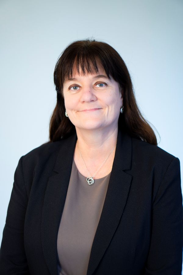 A woman with brown hair wearing a black jacket and brown top smiling warmly at the camera.