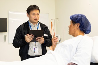 An image of a male interpreter chatting to a patient lying in a hospital bed.