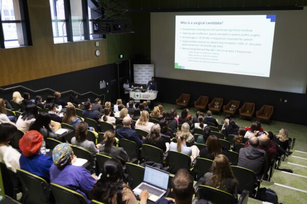 Two people standing at a lectern in front of an auditorium full of people. A slide titled 'Who is a surgical candidate' sits on the screen behind them.
