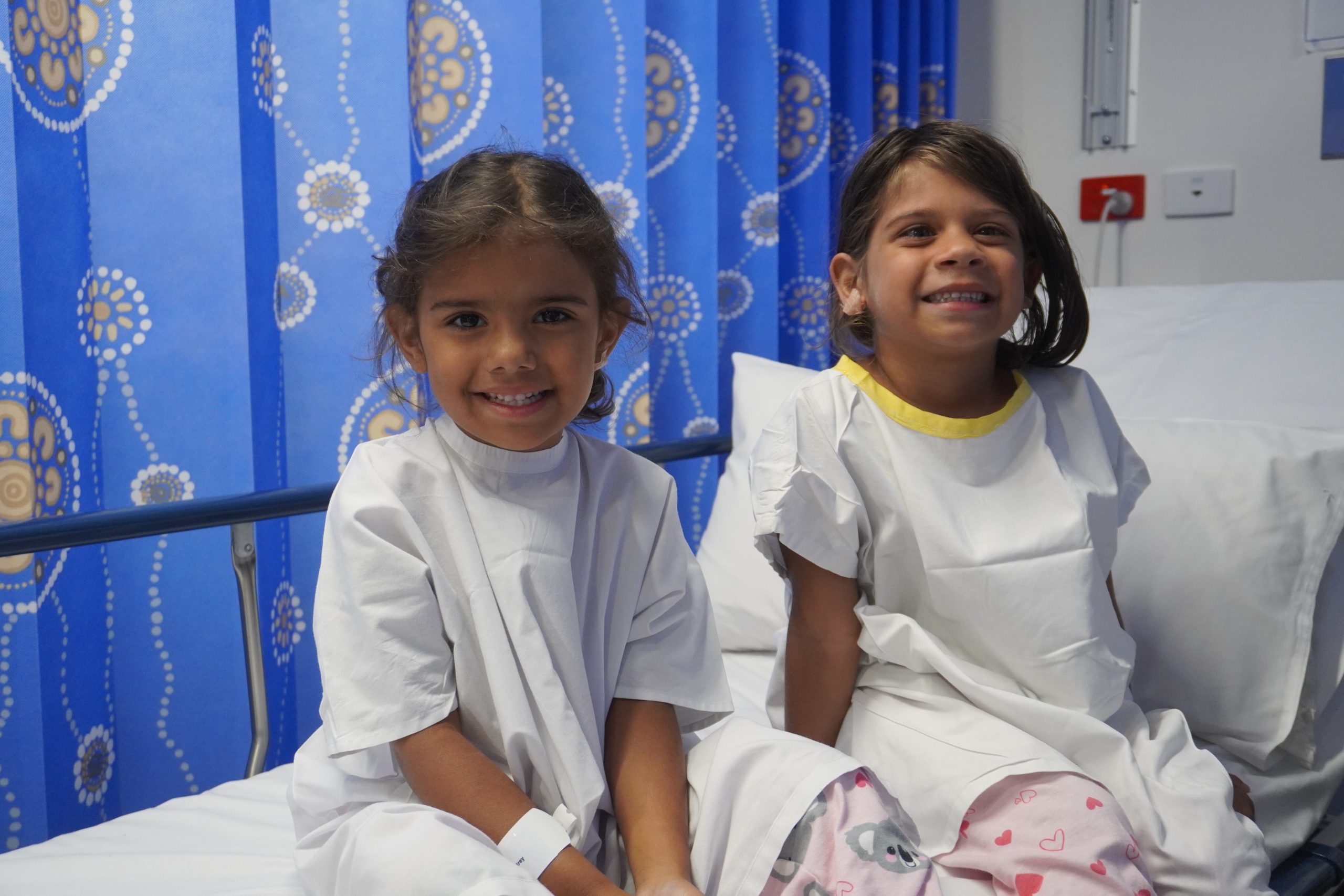 Two four-year-old girls, Audrey and Dianne, sitting on a hospital bed in surgical gowns smiling at the camera against a blue curtain with aboriginal art printed on them.