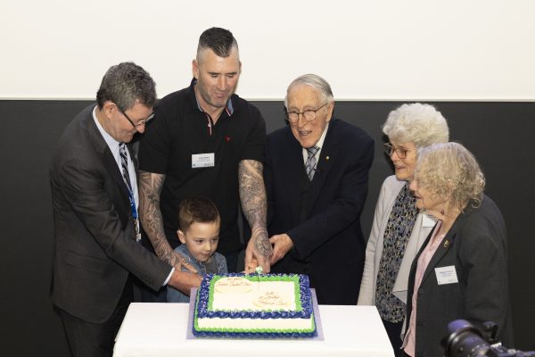 Six people standing around a white, blue and green cake. Several hands are holding the knife as it's being cut into.