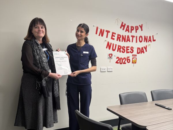 An older woman with brown hair wearing a Brown top, scarf and greay skirts hands over a certificate to a young woman in blue scrubs. Both are smiling against a white wall with a sign hanging which says 'Happy International Nurses Day 2026.
