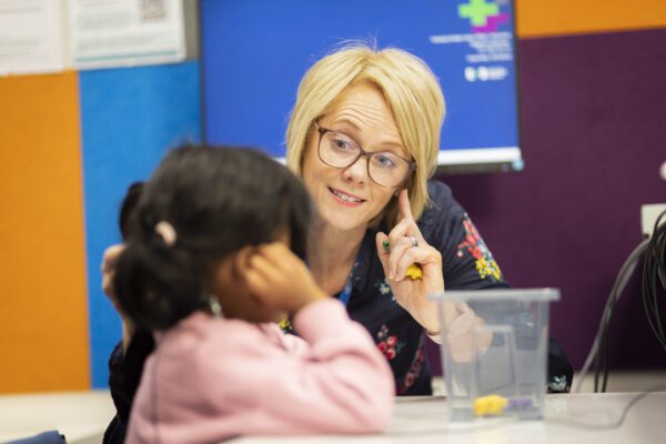 A woman with short blond hair and glasses pointing to her ear as she faces a young girl in a pink jumper in a clinical room with colourful tiles on the wall.