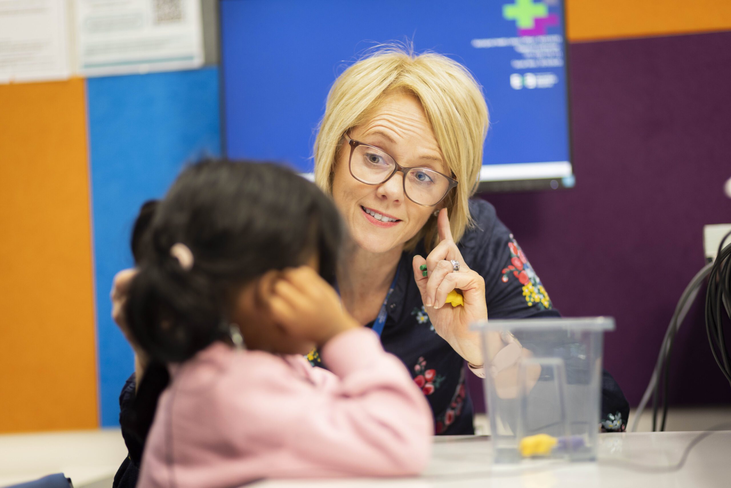 A woman with short blond hair and glasses pointing to her ear as she faces a young girl in a pink jumper in a clinical room with colourful tiles on the wall.