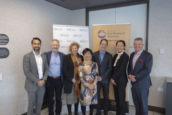 Seven people dressed professionally stand together smiling in front of two banners. ONe banner with the Eye and Ear logo and the second banner with the Centre for Eye Research Australia's logo