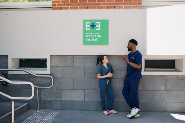 A woman and a man in bllue surgical scrubs leans againt an exterior wall with a large green sign with the Eye and Ear logo.