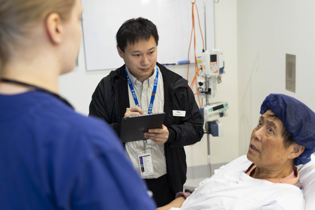 Three people in a hospital setting. A pAtient lying in a bed with a surgical cap on speaks to a medical professional out of focus with back to camera. An interpeter in a black jacket and striped shirt looks and listens to the patient.
