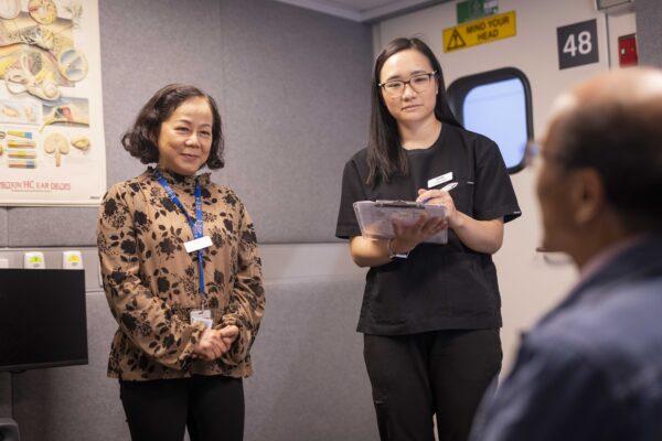 Two women and a man in an audiology booth. The woman on the left is interpreting information from the woman on the right to a man sitting down facing away from the camera.