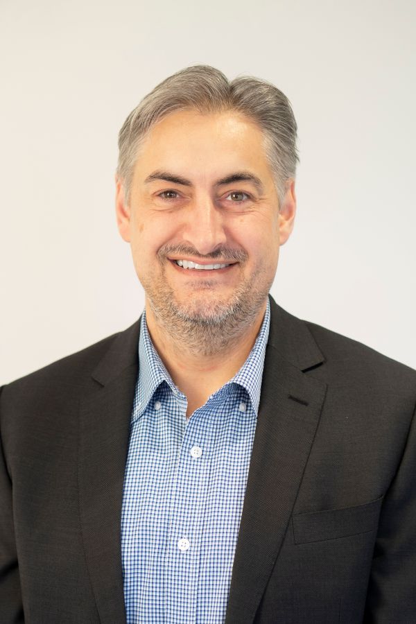 A man wearing a blue and white checkered shirt and black blazer smiles warmly.