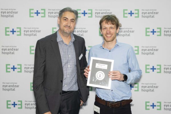 Administrative Excellence Award winner Joachim stands to the right of Executive Director Financial Services and Chief Financial Officer Andre Van Baalen whilst holding is award plaque against a white background with Eye and Ear Logos.
