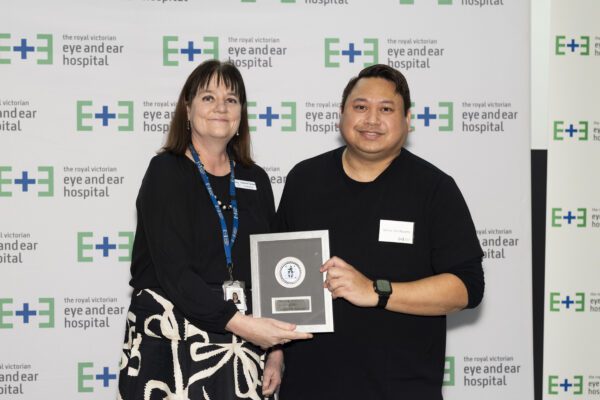 Nursing Excellence Award recipient James stands off to the right of Executive Director Operations and Chief Nursing Officer Leanne Turner with his award plaque against a white backdrop with Eye and Ear logos.