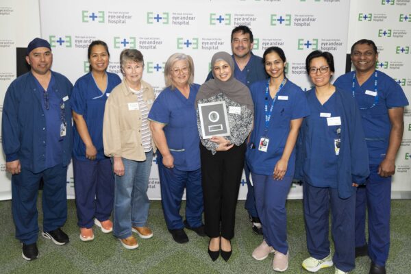 Nine people standing together, most in blue scrubs in front of a white banner with the eye and ear logo holding their award.