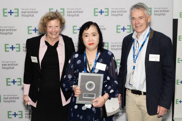 Volunteer excellence award winner Ha stands between Board Chair, Melanie Eagle, and Board Director Bruce Mildenhall holding her award plaque against a white background with Eye and Ear logos.