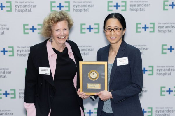 Board Chair Recipient Dr Elsie Chan stands to the right of Board Chair Melanie Eagle holding an award plaque in front of a white banner with Eye and Ear logos on it.