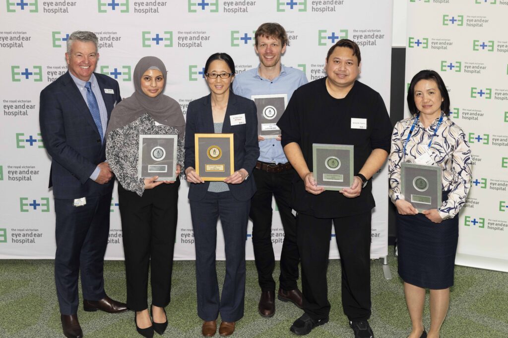 Six people standing together smiling in front of a white banner with eye and ear logos across it. Five of those are holding and award plaque.