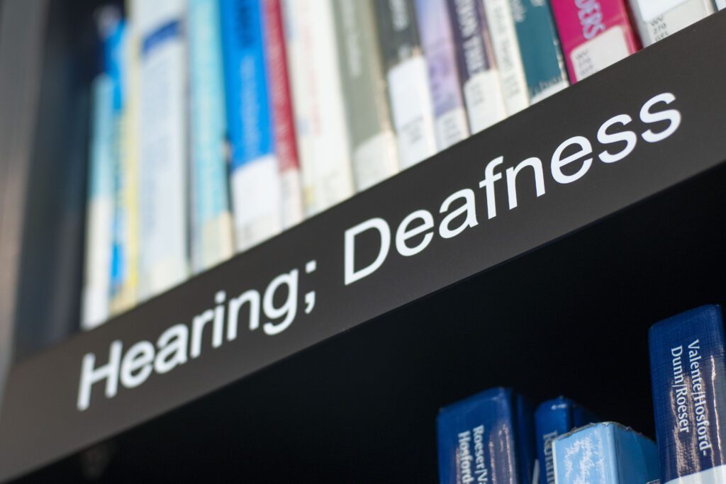 A photo looking up at books on a shelf with a plaque 'Hearing; Deafness' underneath.