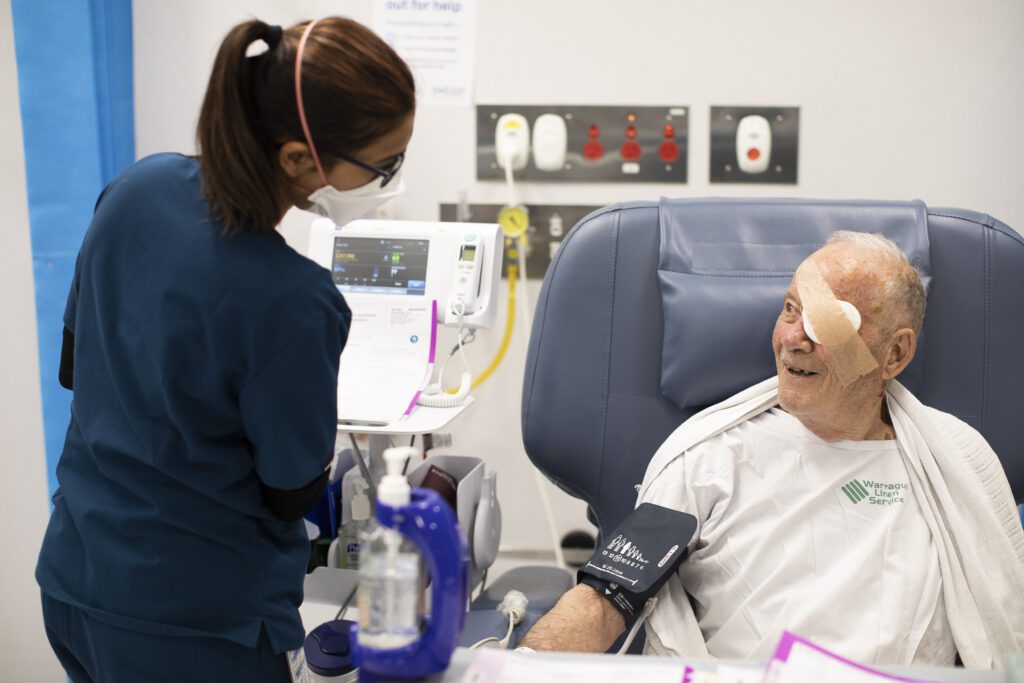 An elderly man sitting in a chair in a hospital gown with an eye patch smiling up at a nurse.